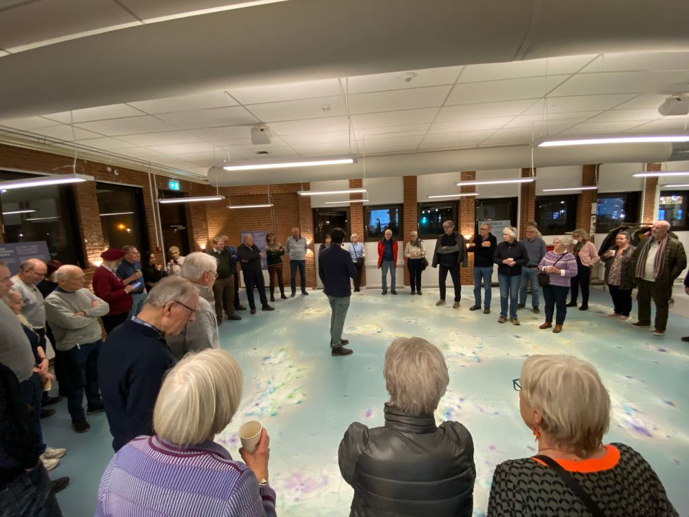 Ca 50 people standing in a circle on a vinyl floor that shows a large scale data visualisation. The people are standing around the edges of the vinyl carpet and look at the person presenting the map in the Center of the circle.