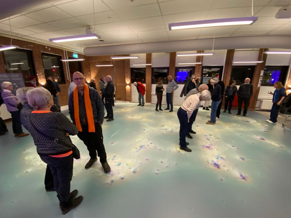 People standing scattered around a room looking on the floor. On the floor there is a large scale data visualisation showing a „map“ of scientific articles about algorithms and AI. 