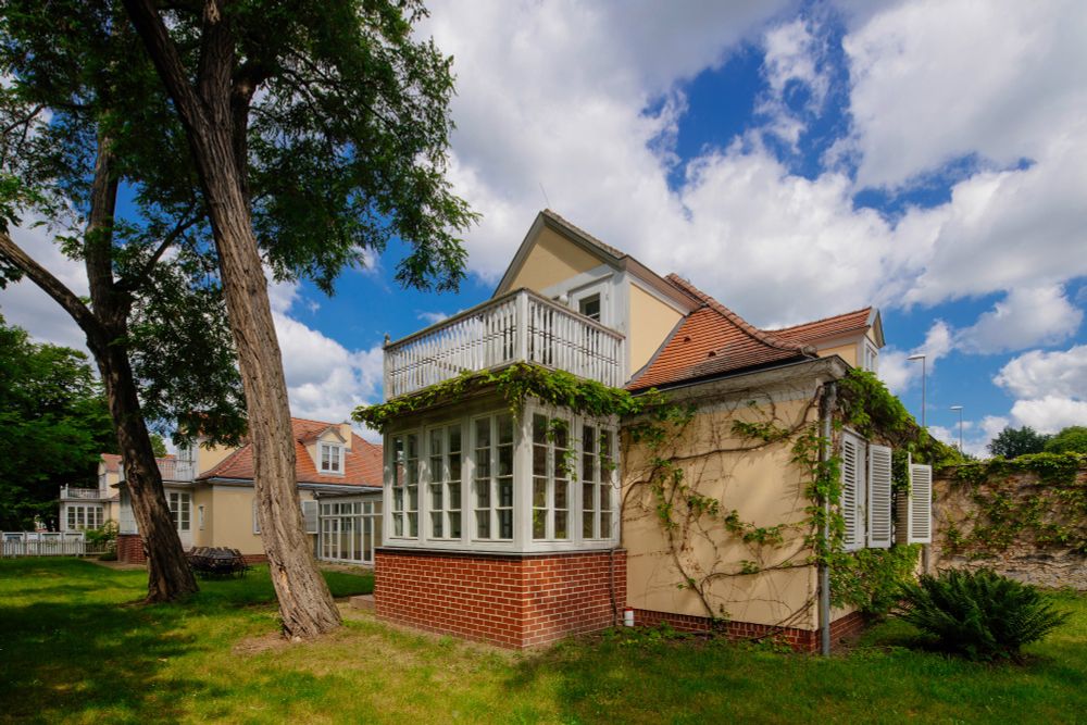 Ein gelbes Haus mit einer Dachgaube, weißen Fensterläden und einem gläsernen Wintergarten wird von Ranken und einer Ziegelsteinverzierung gerahmt.