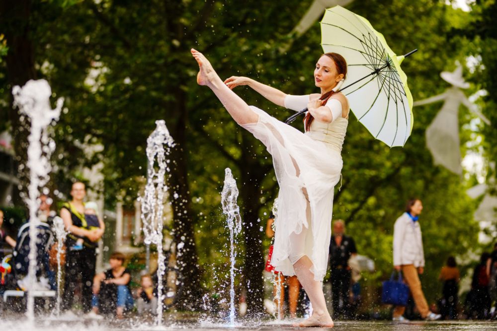 Eine Tänzerin in einem weißen Kleid mit einem hellgrünen Regenschirm tritt in der Nähe von Wasserdüsen in einer Außenanlage auf. Sie balanciert auf einem Bein und streckt ihr anderes Bein hoch.
