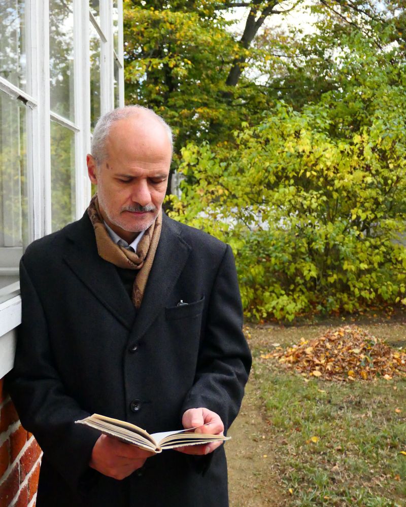 Ein Mann in einem dunklen Mantel und mit Schal steht draußen an einer Hauswand und liest ein Buch, im Hintergrund Bäume und Laub auf dem Boden.