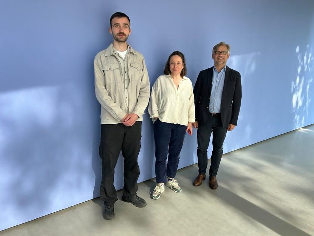 Drei Personen stehen nebeneinander vor einer einfarbigen, hellblauen Wand in einem gut beleuchteten Raum mit glattem Betonboden. Von links nach rechts: Ein junger Mann mit kurzem Bart, hellgrauer Hemdjacke und dunkler Hose, eine Frau mit schulterlangen braunen Haaren, weißer Bluse, dunkelblauer Hose und Sportschuhen, sowie ein älterer Mann mit grauem Haar, Brille, dunklem Sakko, hellblauem Hemd und Jeans. Alle drei blicken freundlich in die Kamera.