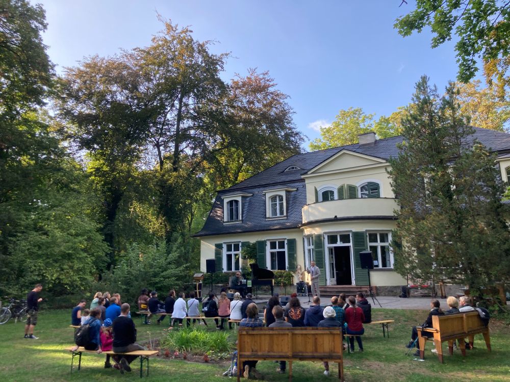 Mehrere Dutzend Menschen sitzen auf Bänken und Holzmöbeln im Garten vor einer Villa mit grünen Fensterläden. Sie verfolgen eine Veranstaltung auf einer kleinen Bühne mit Mikrofon und Flügel. Die Szene spielt bei sonnigem Wetter unter hohen Bäumen, die das Gelände umgeben. Die Stimmung ist ruhig und konzentriert.