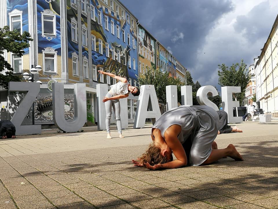 Eine Tänzerin in grauem Kleid kniet auf einem gepflasterten Platz, ihr Kopf ist auf den Boden gestützt. Hinter ihr ist das Wort ZUHAUSE in großen grauen Betonbuchstaben aufgestellt.