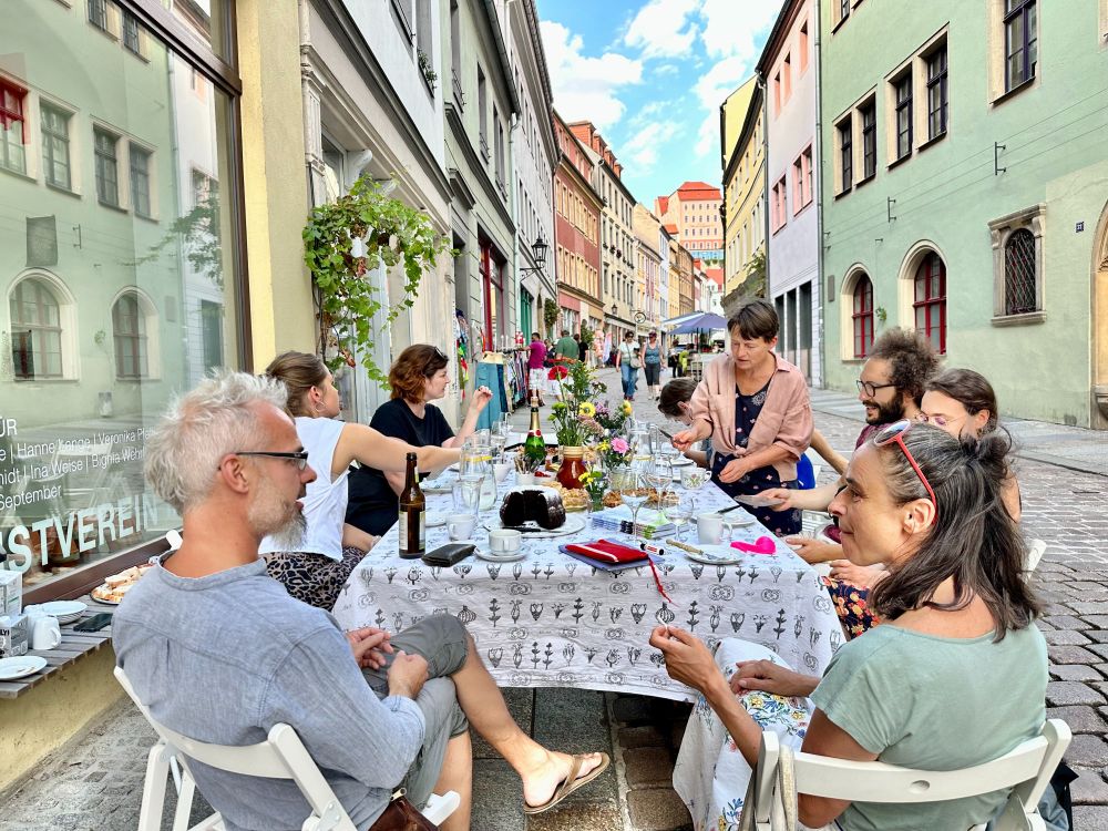 Eine Gruppe von Menschen sitzt an einem langen, festlich gedeckten Tisch auf einer gepflasterten Altstadtstraße. Sie unterhalten sich, essen und trinken gemeinsam. Der Tisch ist mit Blumen, Gläsern und Geschirr geschmückt. Im Hintergrund sind bunte historische Häuser und Passanten zu sehen. Die Atmosphäre wirkt sommerlich und gesellig.