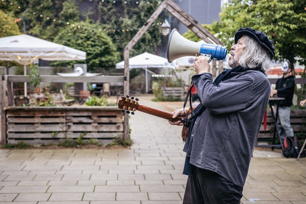 Ein Mann mit einer umgehängten Gitarre steht im Freien und ruft durch ein Megaphone. Im Hintergrund ist ein Biergarten mit Sonnenschirmen und Bäume zu erkennen. 