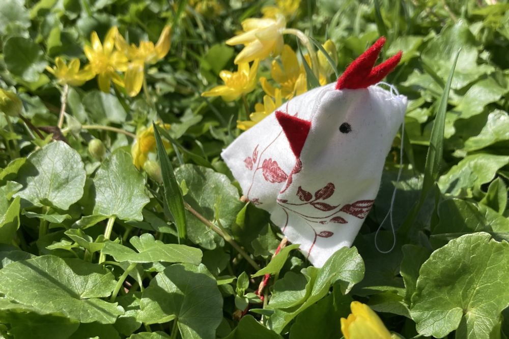 Ein kleines, handgenähtes Stoffhuhn mit rotem Kamm und Schnabel sitzt auf einer grünen Wiese mit gelben Frühlingsblumen. Das Huhn besteht aus weißem Stoff mit einem roten floralen Muster.