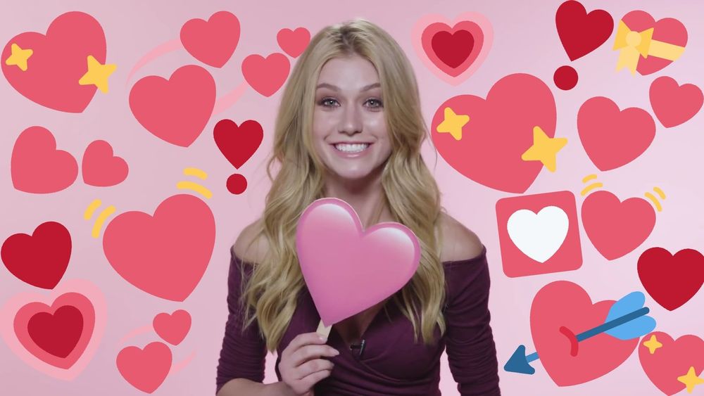 Kat McNamara, very happy, holding a heart-sign and surrounded by hearts