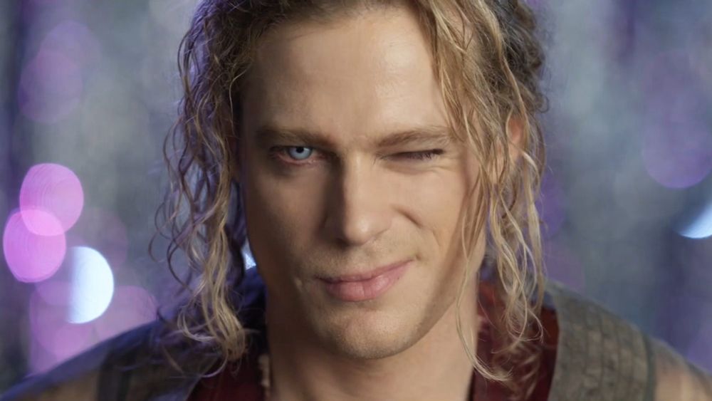 Headshot of Sam Reid as the Vampire Lestat with his blond hair in a bun( I think) some stray curls escaping to frame his face . Winking at the audience 