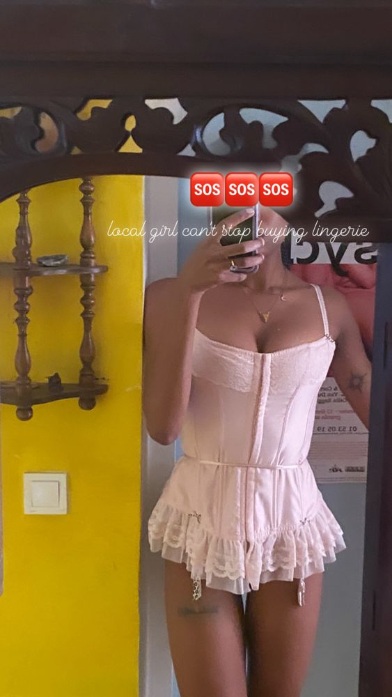 A mirror selfie of OP (Aura) wearing a pink corset and bare legs. The wall next to her is yellow, and written over her face is a text that says "SOS: Local girl can't stop buying lingerie"