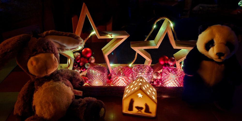 Ein Plüsch-Esel und ein Plüsch-Panda sitzen an den Enden einer Weihnachtsdekoration mit großen Sternen und vier brennenden Kerzen, im Vordergrund ist eine kleine, beleuchtete Krippe aus weißer Keramik zu sehen. 