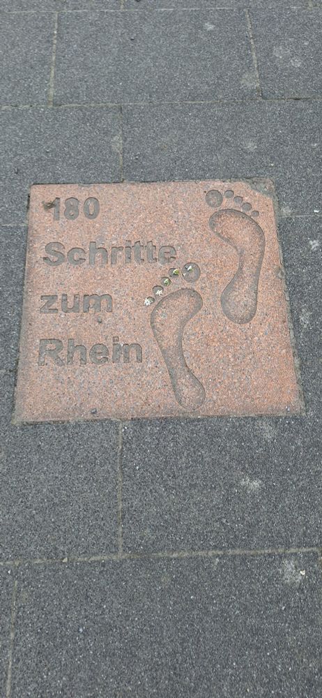Eine im Boden eingelassene Platte mit zwei Fußabdrücken und der Aufschrift "180 Schritte zum Rhein"