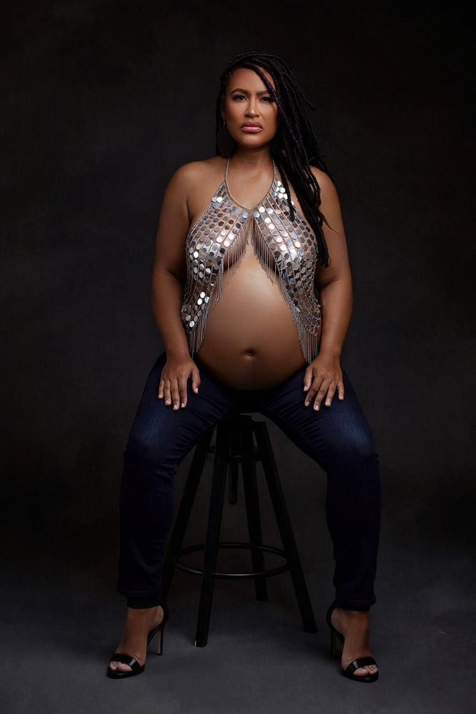 Maternity photo: pregnant Black woman seated on a stool looking directly ahead, hands resting on her thighs. She's wearing dark wash skinny jeans, a metallic halter top that covers her breasts and drapes down to frame her belly. 