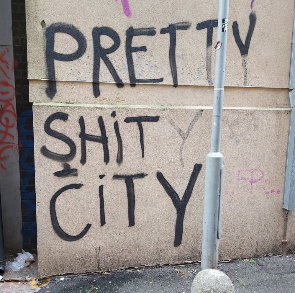 A beige wall with graffiti which originally read 'pretty shit city'. Someone has then tried to correct it by adding just one 'y' after 'shit'