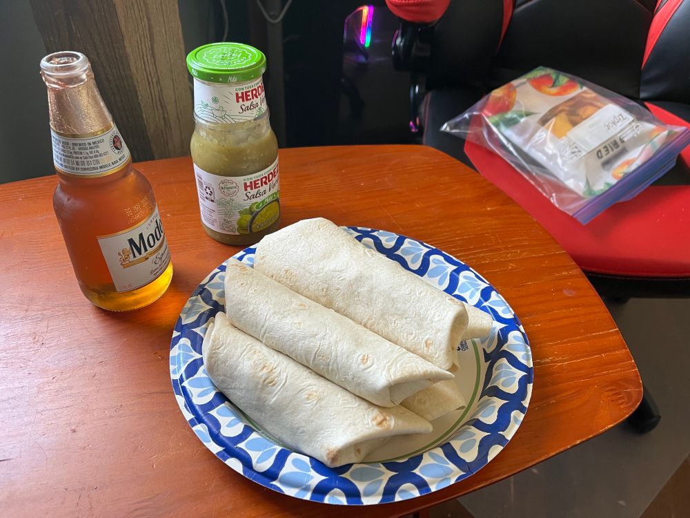 Paper plate with a stack of burritos on a TV tray next to a bottle of salsa verde, an open Mexican beer, and a bag of dried mango leather