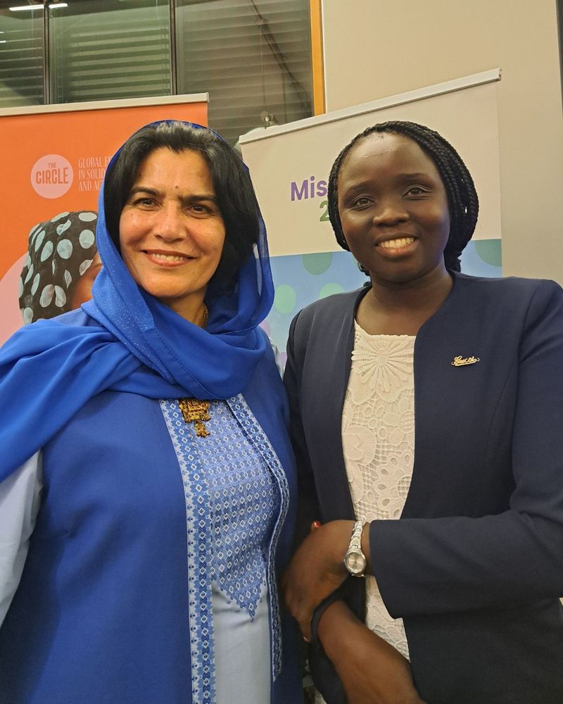 Photo of a woman dressed in a blue outfit with a blue shawl over her head, and a woman in a white dress and blue blazer, smiling towards the camera