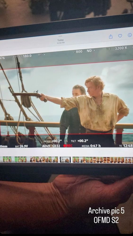an archive pic of ofmd s2. rhys darby, playing stede bonnet, is holding a gun up to shoot at something off screen. he has his left hand on his hip