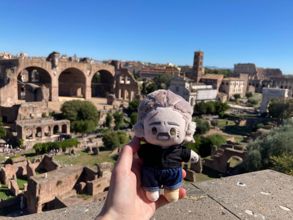 rights izzy doll held up over the roman forum. to the left is the basilica of maxentius, to the right santa francesca romana and the colosseum. 