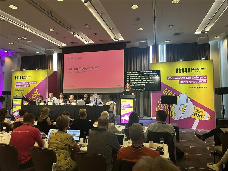 Photo of Alison McGovern speaking from a lectern on the right side of the stage. Behind her to her left is a red screen that says, "Alison McGovern MP". Immediately behind her is a screen with live captioning of her speech. To her right is yellow and pink staging that says "M U Musicians' Union, Welcome to Delegate Conference 2025 #MUConference". Alison is in the centre. To her right is the top table with six people listening, and in the foreground are members sitting at rows of tables