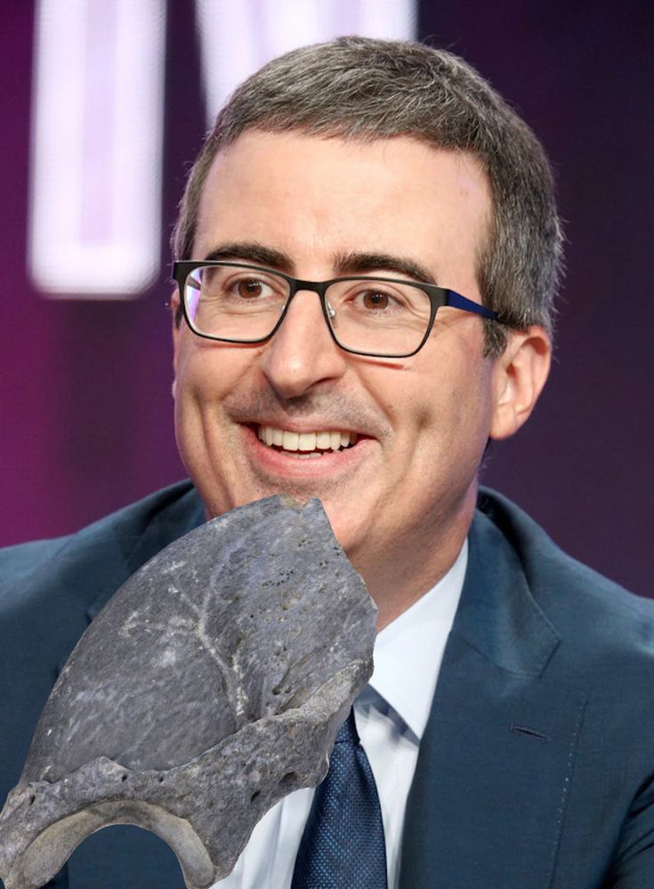 John Oliver smiling in a blue suit and blue tie with a dark sloth claw fossil brought to his chin