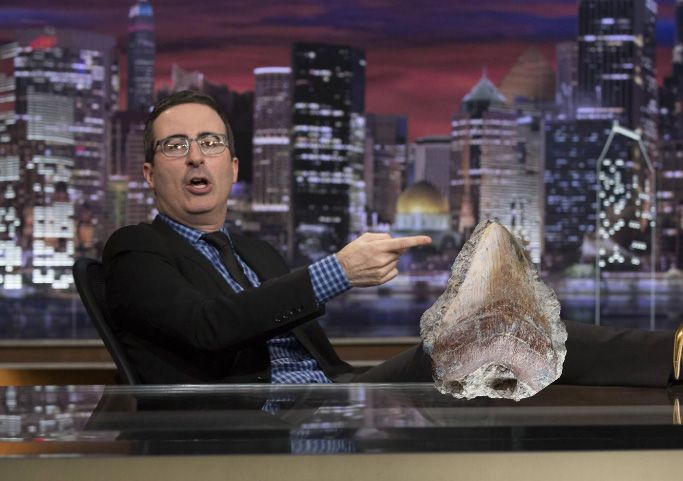John Oliver with his feet up on his desk pointing to a large fossil shark tooth in a small amount of matrix rock on his desk