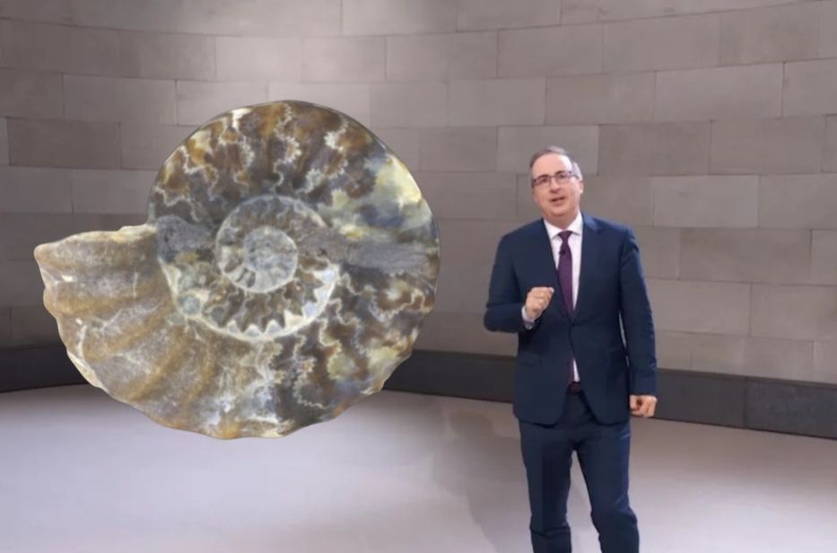 John oliver stands in a dark suit in a museum gallery like space, standing next to a fossil ammonite cut in half to show complex sutures
