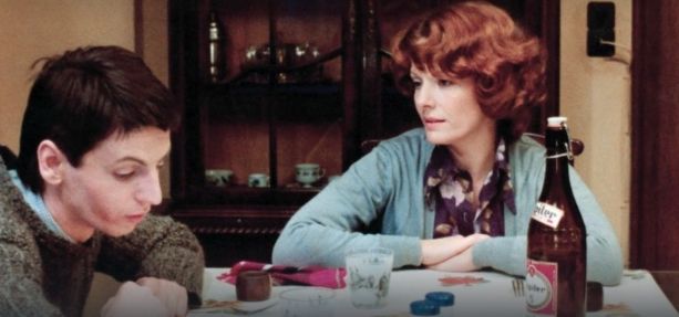 Still from Jeanne Dielman: a red headed white woman with curly hair regards a brown haired boy at a white covered table. She looks like she wants to tell him something, he is not looking at her