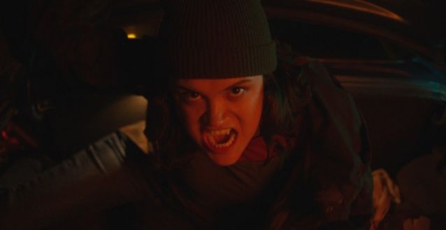 Pale dark haired teenage girl in a beanie baring her elongated canines at the camera