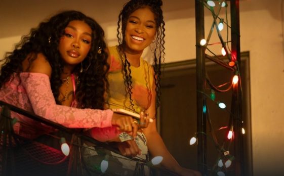 Still from one of them days: Sza (pursed lips) and keke Palmer (smiling widely) looking down from a fairy light adorned metal balcony