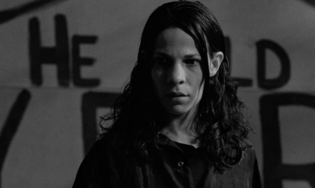 Black and white shit. White woman with matting dark hair in front of a wall with words on it ('He') is visible. She is looking in confusion at something offscreen