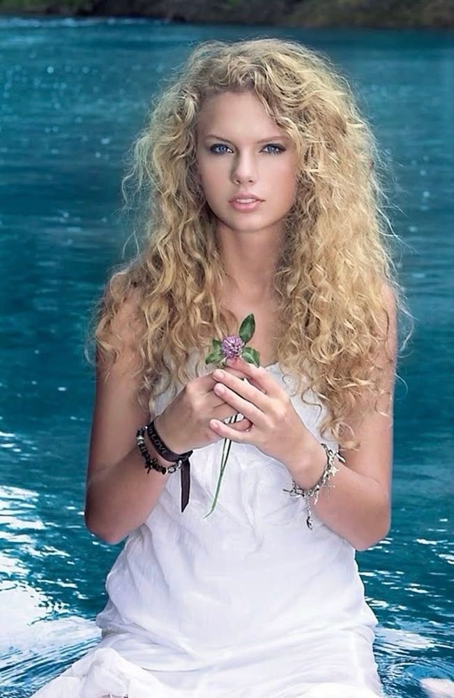 taylor swift during her debut era sitting in a pond with a flower in her hand wearing a white dress and curly blonde hair