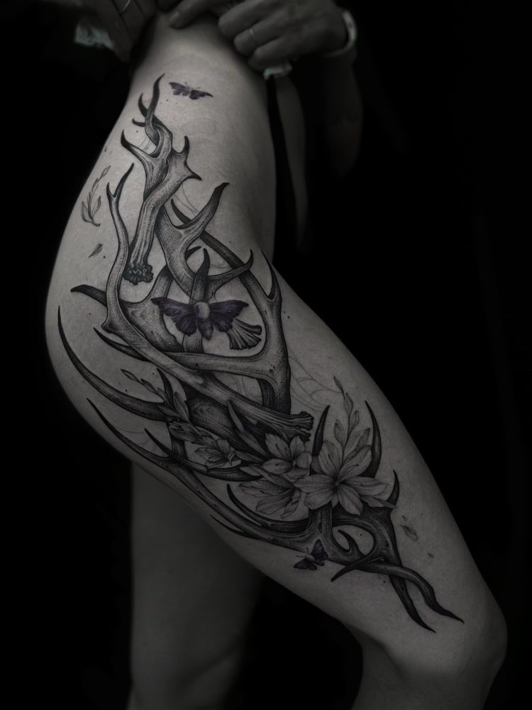 A photo of a woman’s side thigh with a big tattoo, on a black background. Tattoo of an arrangement of antlers, flowers and moths. 