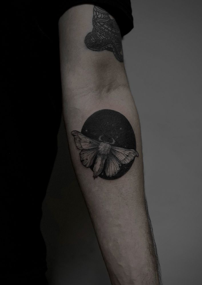 Tattoo of a moth flying over a black hole/elipse, on an inner arm. 