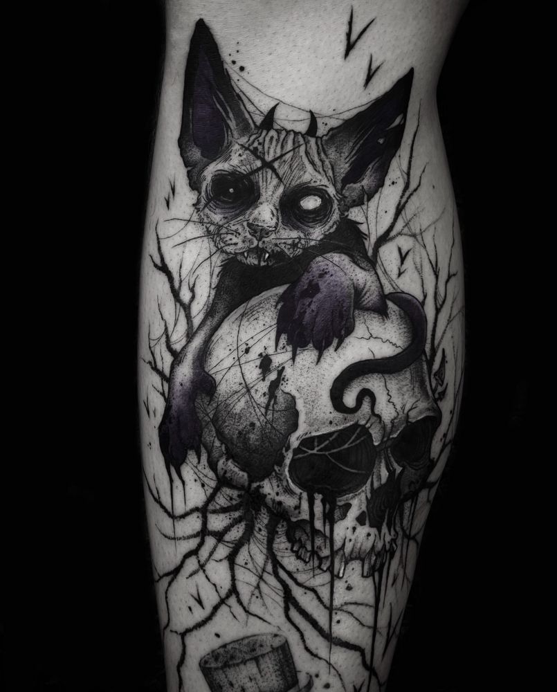 Photo of a tattoo of an undead Devon Rex cat on a human skull, surrounded by bats, branches and spiderwebs. 