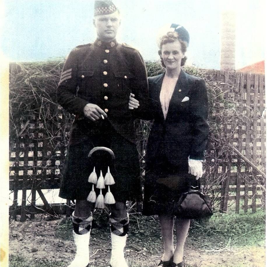 A man in a Canadian army uniform that features a Scottish kilt stands next to his wife. The picture was taken sometime during WWII, which you can tell by their clothing styles.
