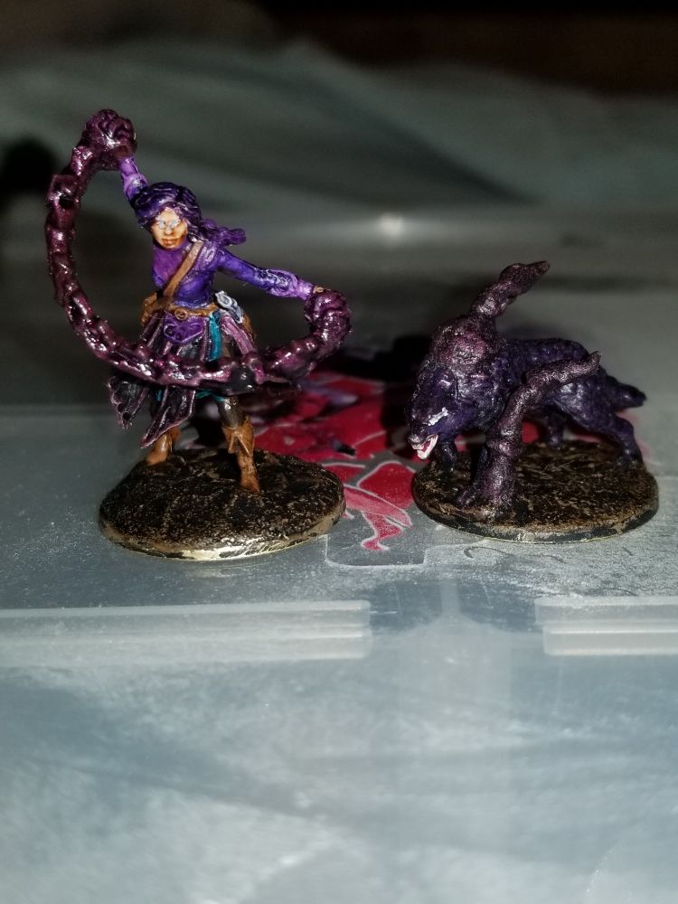 Two miniatures, one of a purple clad human with a dark colored magic effect going from the hands. And the second miniature is a dark purple wolf with wispy smoke like effects coming off of it.