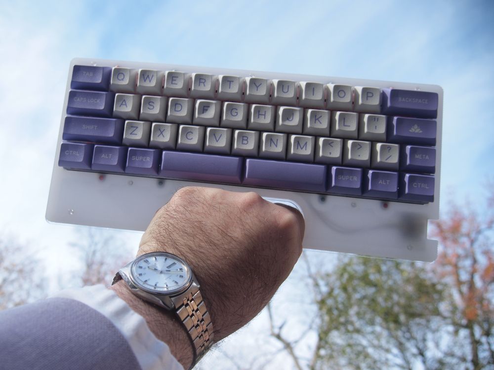 White stacked acrylic keyboard with pink switches and white and lavender keycaps being held up to the sky, the arm is wearing a lavender jacket, white shirt and silver watch 