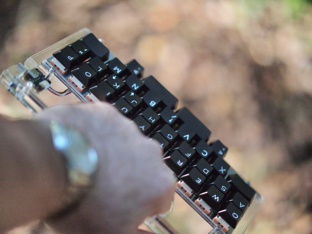 40% keyboard in an acrylic case featuring a handle. It is being held by the handle and the photo is taken from the perspective of the holder. 