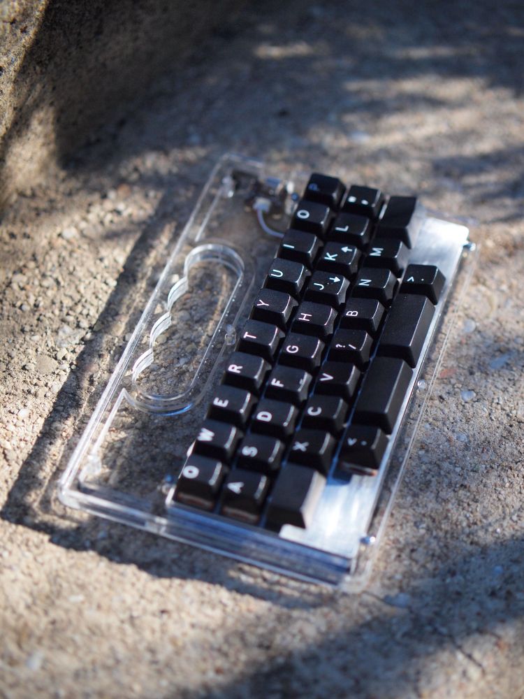 40% keyboard in an acrylic case featuring a handle. It is sitting on a warm gray stone staircase. 