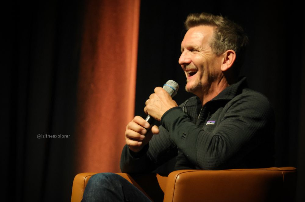 Sebastian sitting in an arm chair on stage. He is holding a mic in both of his hands and is wearing a big smile on his face 