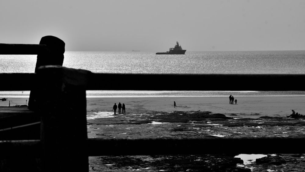 Late afternoon on a French beach opposite Dover.
A barrier on the sea wall frames 3 groups of people on the beach, a child running from one group to another. Behind them, a lifeboat is moored, awaiting a rescue mission in case any refugees attempt to cross to England.