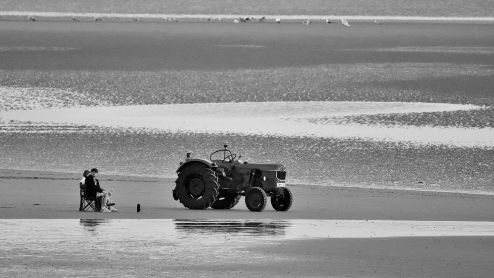 Two people are sitting on folding chairs on a beach. In front of them is a mug and a stationary farm tractor. They seem to be watching the tractor.