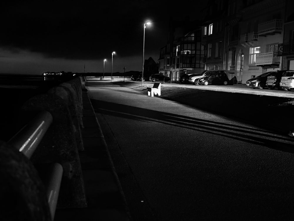 The sea front of Ambleteuse at night. In the distance, the village of Audresselles, on the left, the sea at low tide and on the right, the street. A car passes by, a couple sitting on a bench draws a dark line in the light of the headlights.