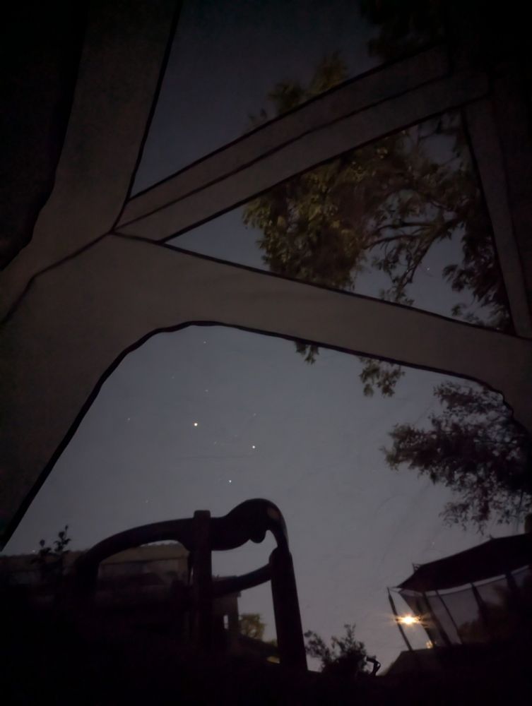A photo of the night sky taken from inside a tent. Some stars shine faintly in a greyish sky, and the silhouettes of trees and children's play equipment fade into the dark edges of the photograph.