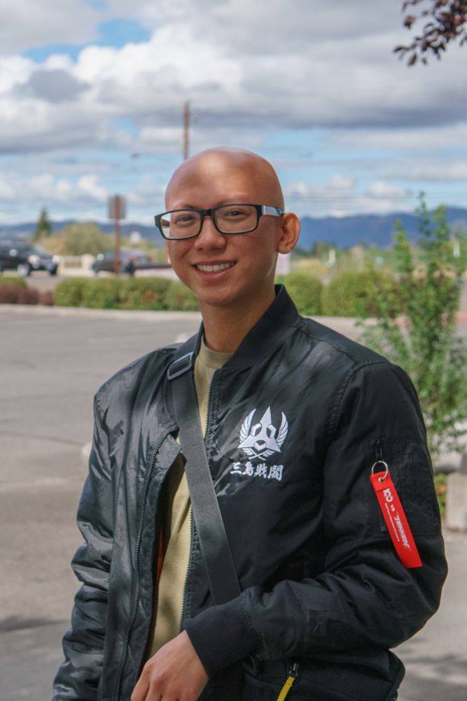 FORMAT: Vertical portrait
SUBJECT: Waist-up of SoCal SUMA with his arm leaning against his camera bag.
BACKGROUND: A blurry parking lot and passing cars. Behind them are the mountains and clouds of Central Point, Oregon.