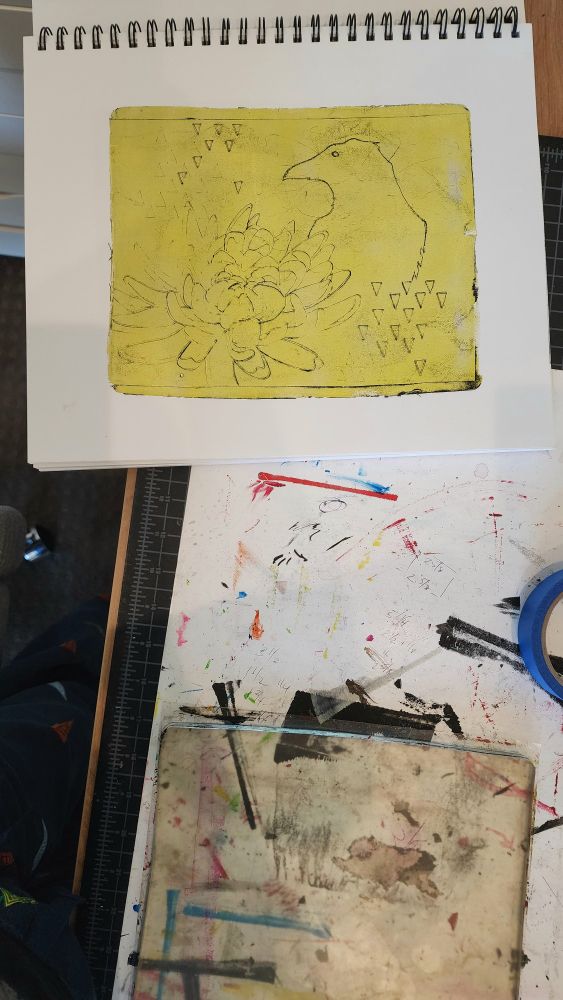 Photo looking down at my studio worktable. At top of image is my sketchbook with a black on yellow monotype print of a crow and a chrysanthemum. There is a white poster board with paint marks all over it underneath a clear gelliplate at the bottom of image.