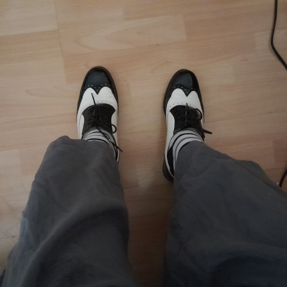 Zwei graue Hosenbeine, schwarzweiß geringelte Socken, schwarzweiße Lackschuhe und ein heller Laminatboden. 