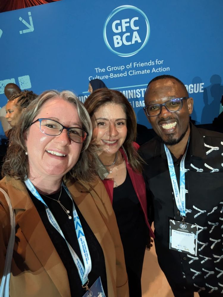 Jenny Hay in a brown jacket, Princess Dana Firas in a red jacket, and Simon Musasizi in a black shirt smile in a selfie in front of the Group of Friends for Culture-based Climate Action logo 