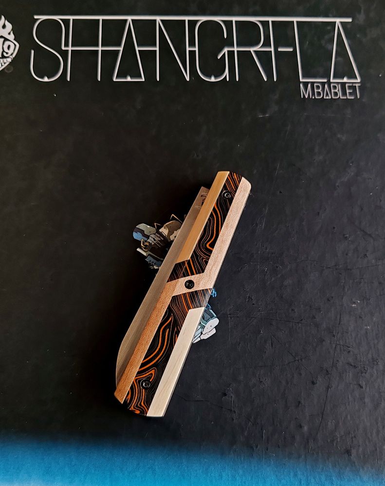 Couteau avec un manche orange et noire avec des formes géométriques crème 
Couteau replié 
En fond l'album shangri-la de m bablet