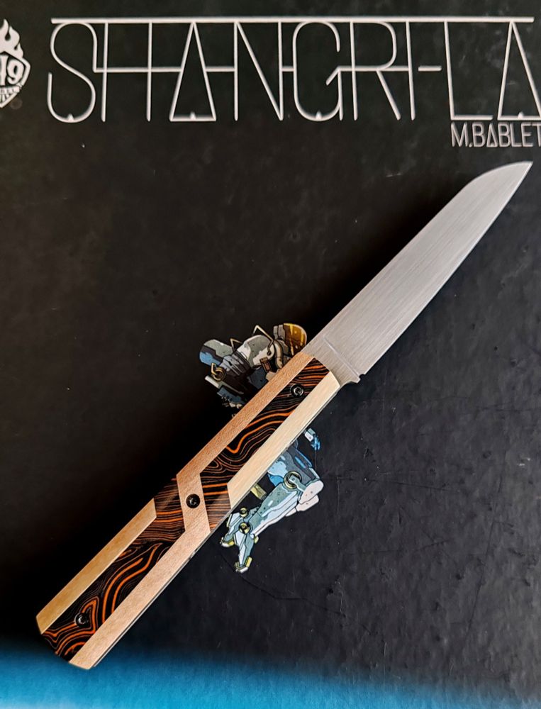 Couteau avec un manche orange et noire avec des formes géométriques crème 
Lame pointé vers le haut à droite
En fond l'album shangri-la de m bablet